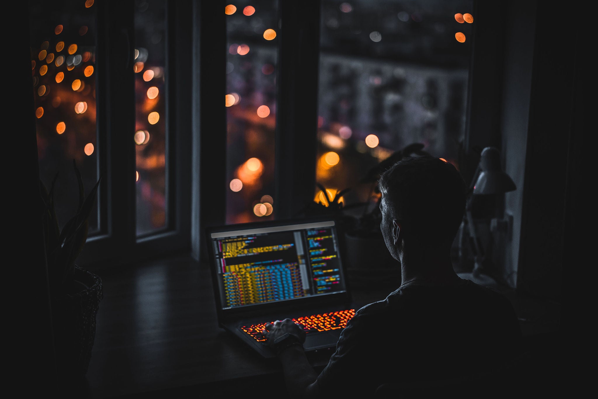 Programmer working in dark room at window with view on city night lights